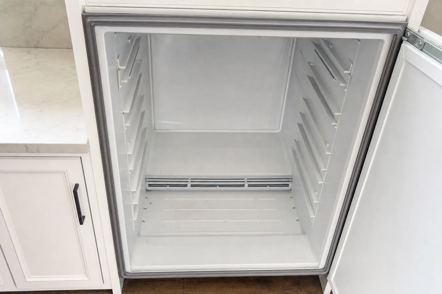 Open built-in refrigerator compartment, emptied out, grey door gasket and shelf rails exposed