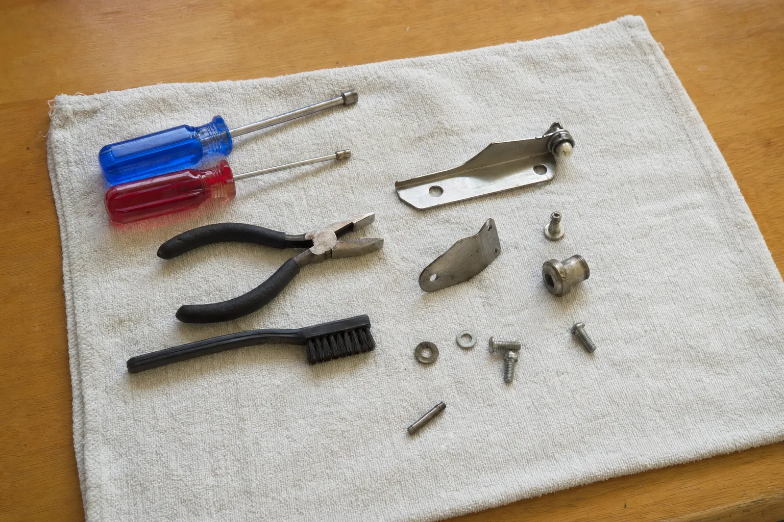 A hinge arm with its roller, a bent bracket, a bushing, washers and a pin on a towel beside two nut drivers, pliers and a nylon brush
