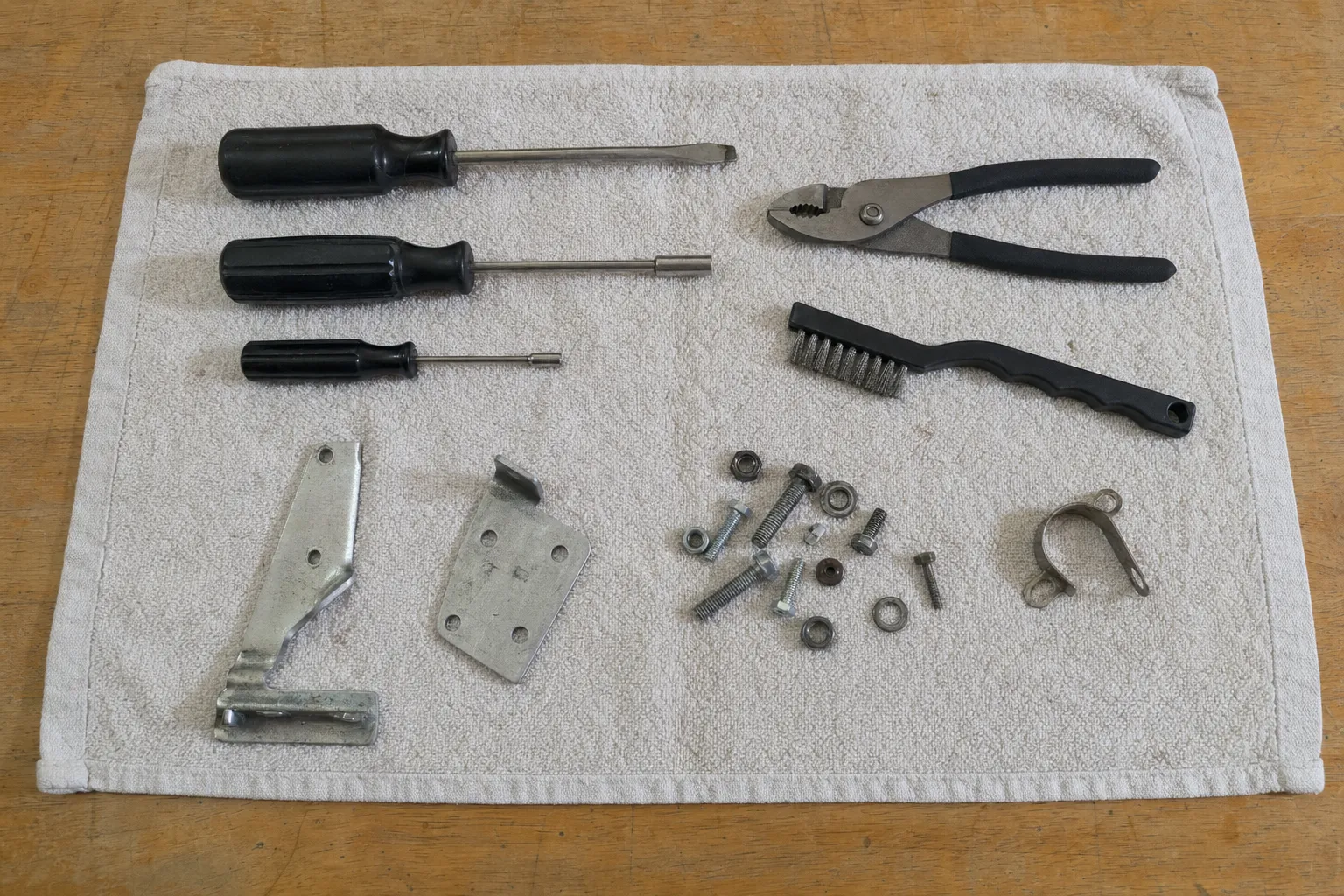 Three black-handled drivers, slip-joint pliers and a wire brush on a towel with two steel plates, a pipe strap, bolts, nuts and washers