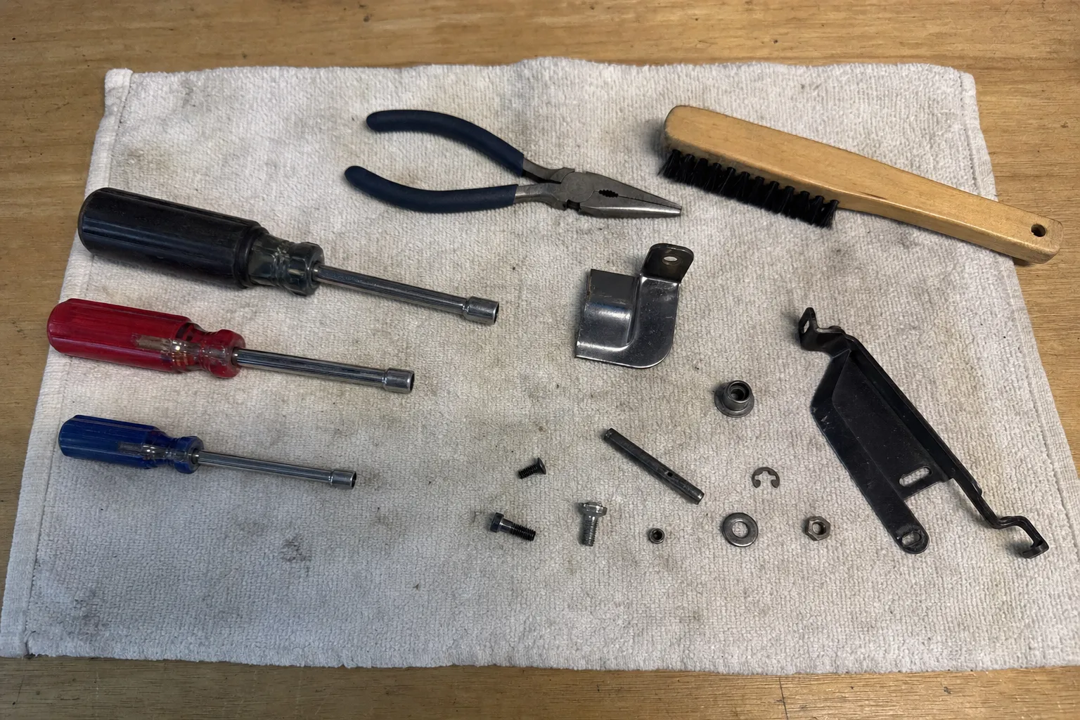 Three nut drivers, long-nose pliers and a wooden brush on a towel with a black steel arm, a bent bracket, a pin, a bushing, a clip and screws